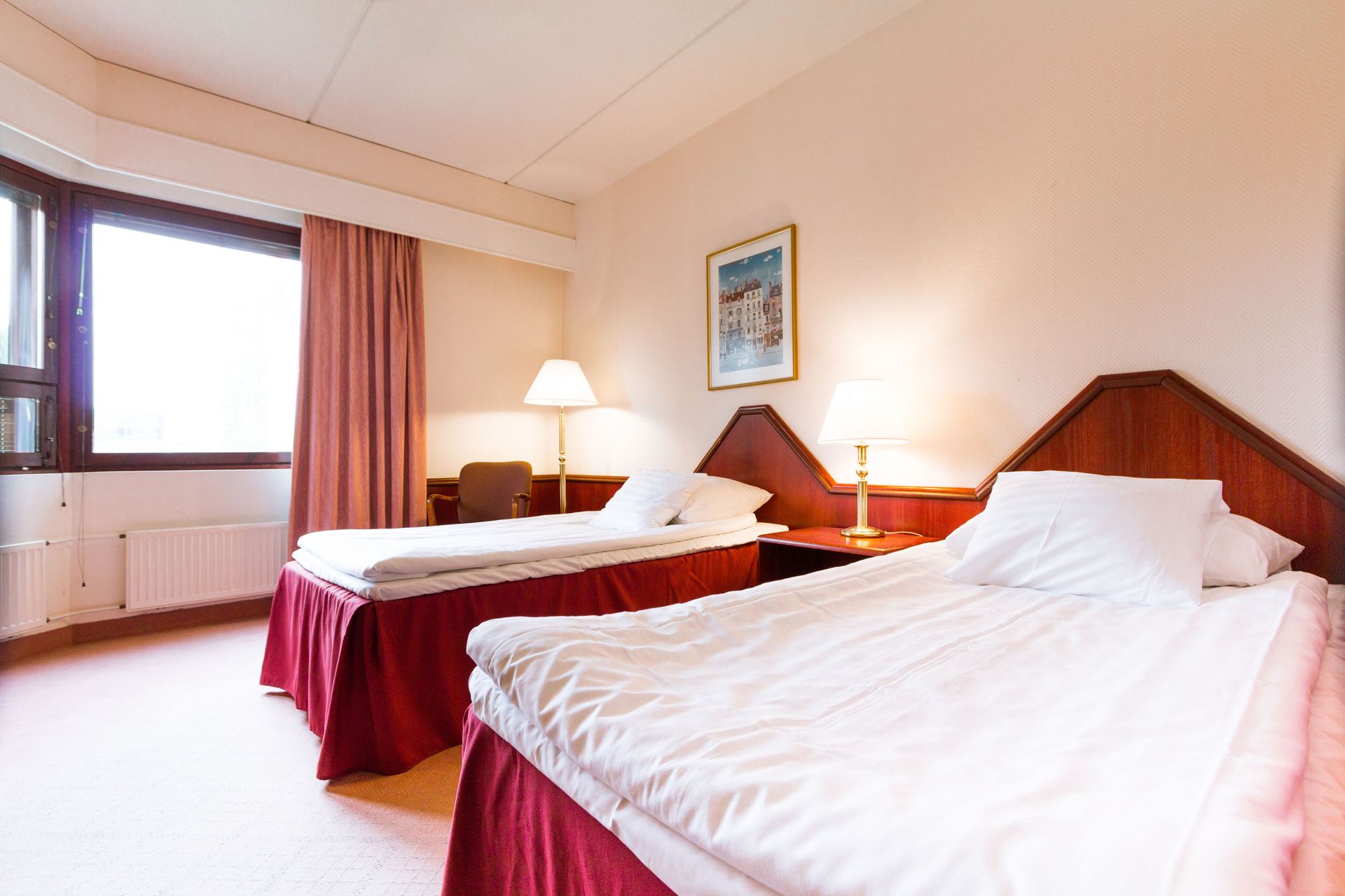 Hotellihuone, jossa on kaksi erillistä sänkyä valkoisilla lakanoilla, punaiset helmat ja ikkuna verhoilla.