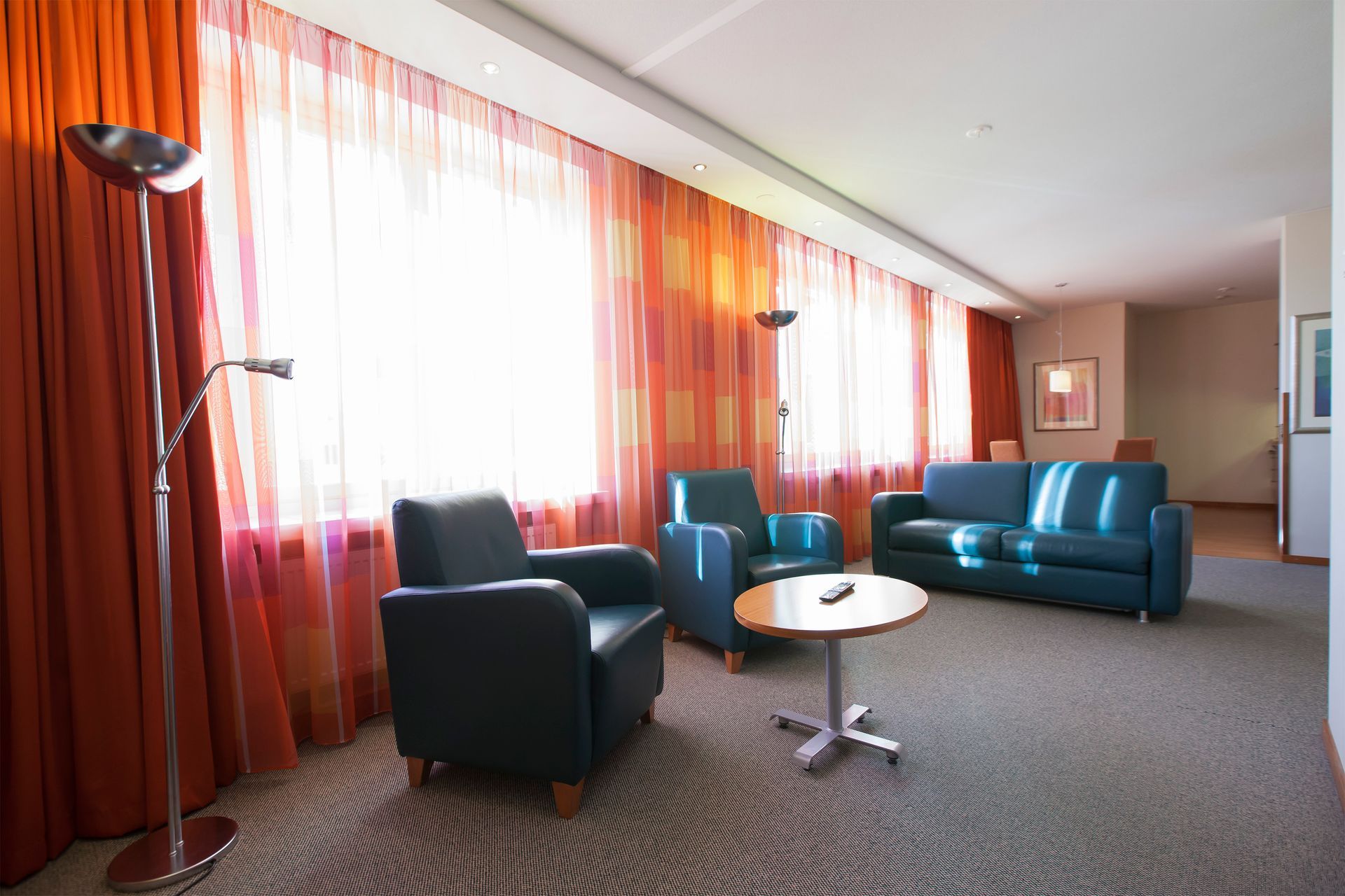 Hotellin oleskelutila, jossa on kaksi tummaa nahkanojatuolia, yhteensopiva sohva, pyöreä sivupöytä ja oranssit verhot.