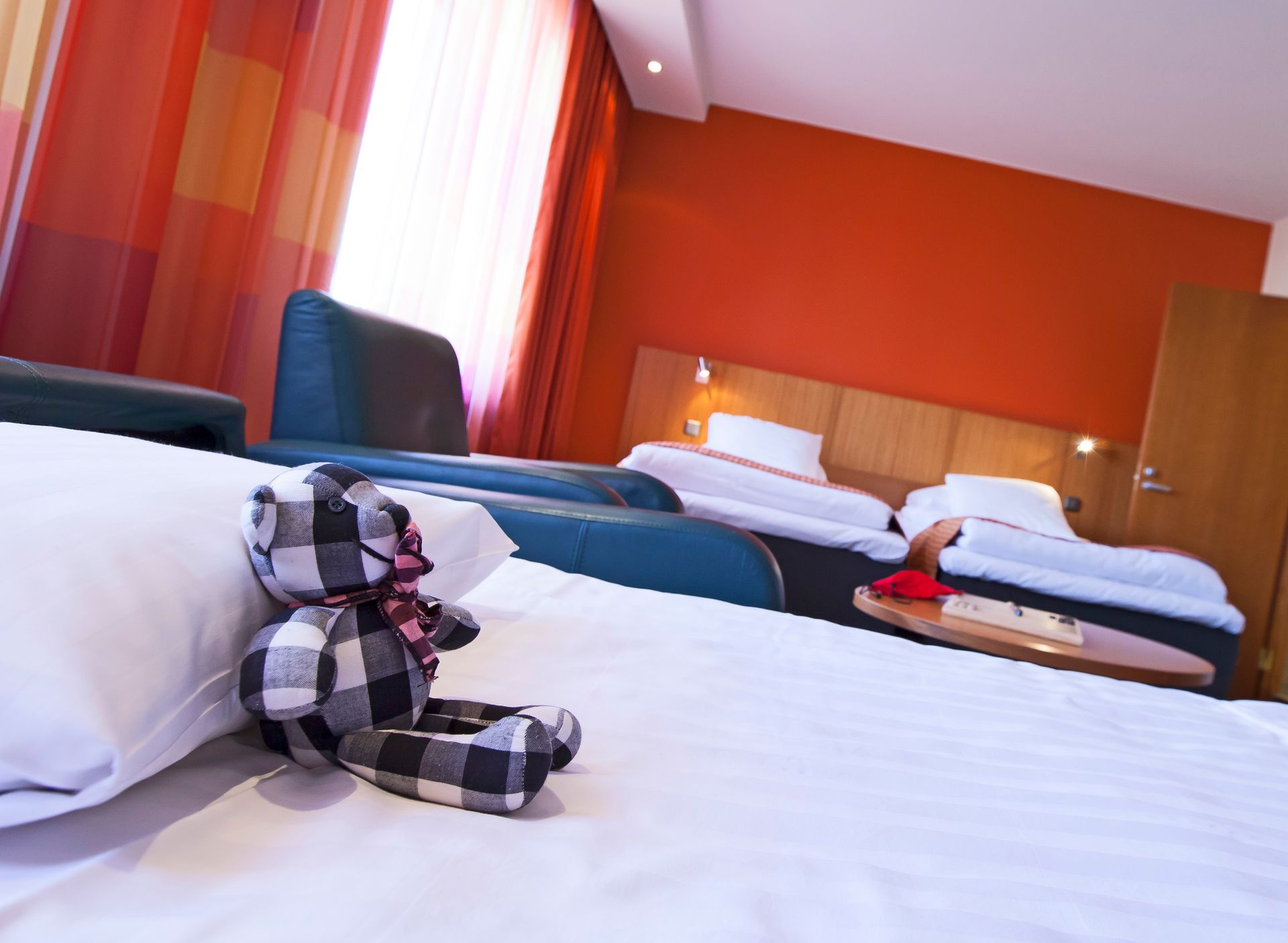 Pehmoinen ruudullinen nallekarhu istuu valkoisella sängyllä hotellihuoneessa, jossa on oranssit seinät ja kaksi lisäsänkyä taustalla.