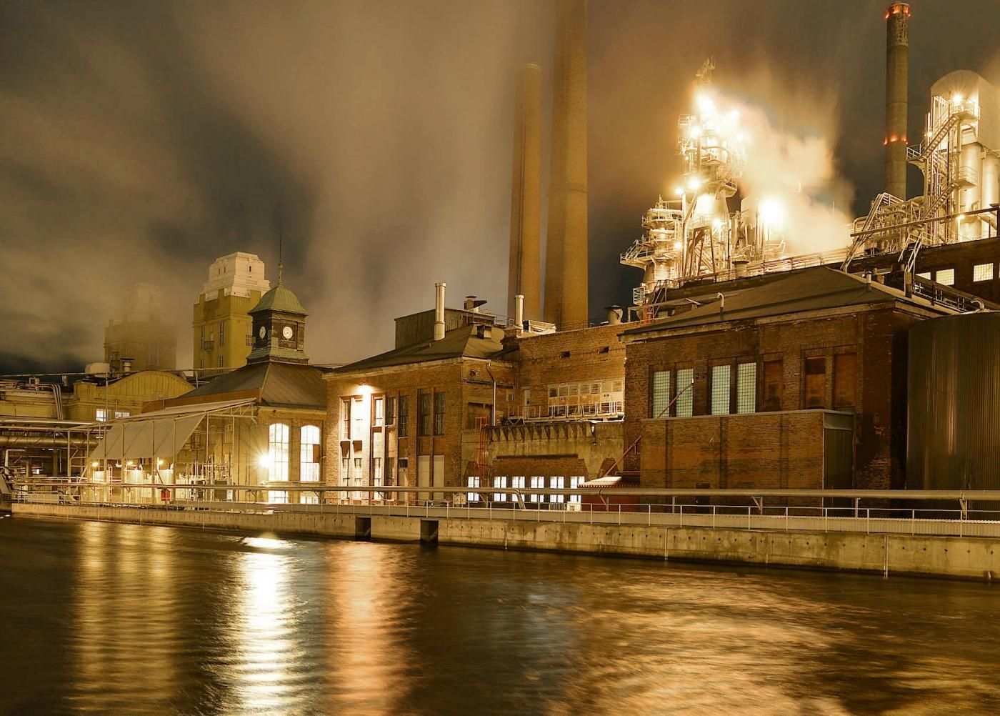 Industrial factory with smoke stacks at night, illuminated by golden lights reflecting on the water in the foreground.