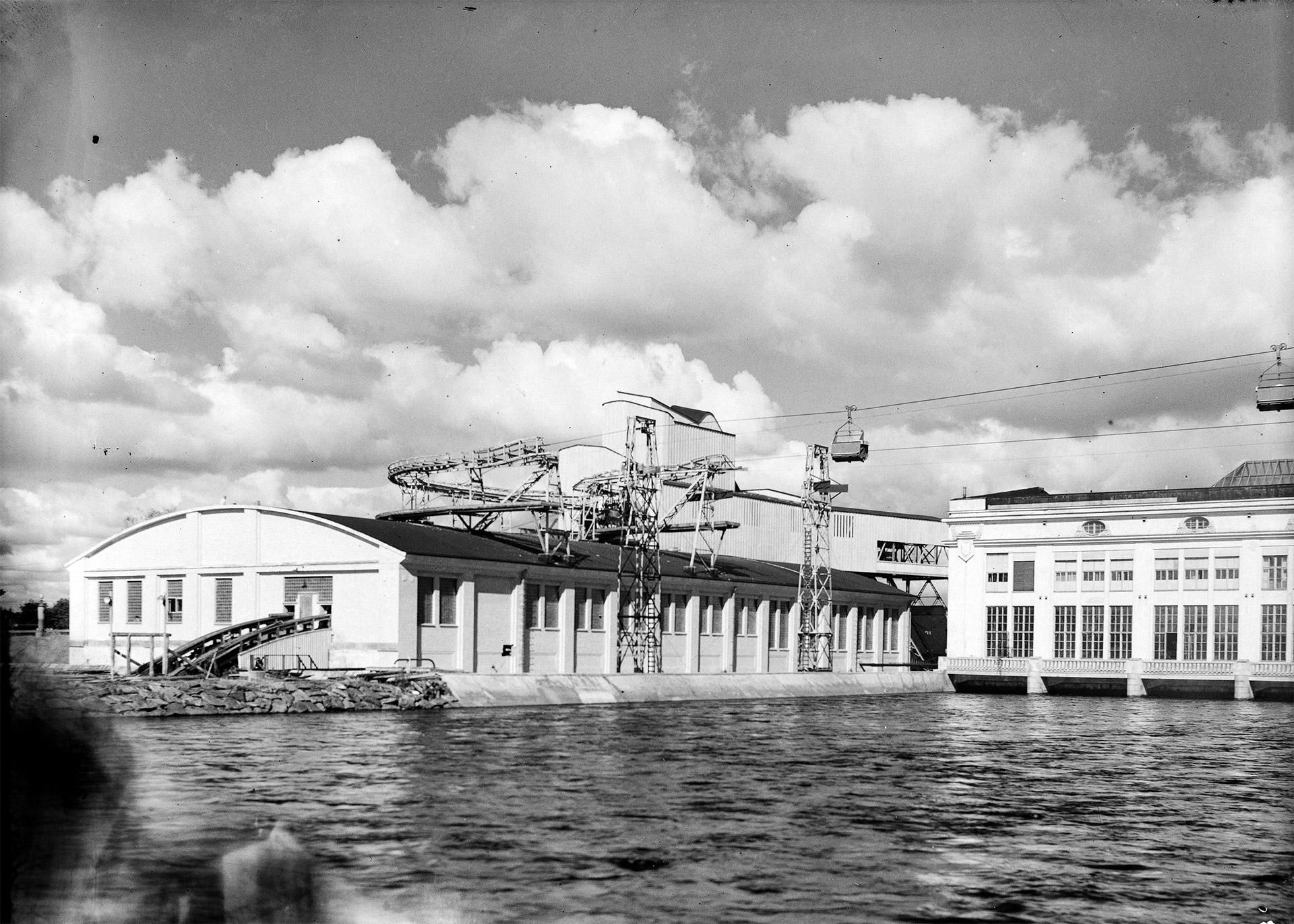 A black-and-white view of a lakeside industrial facility with a cable car system spanning between two buildings.
