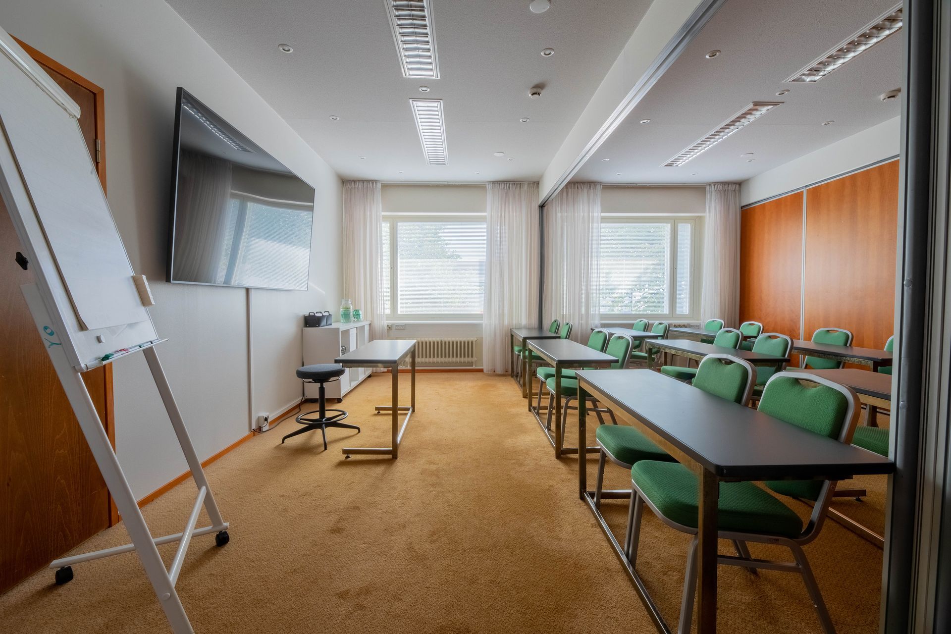 A meeting room with rows of tables, green chairs, a white board, a large mounted screen, and windows with white curtains.