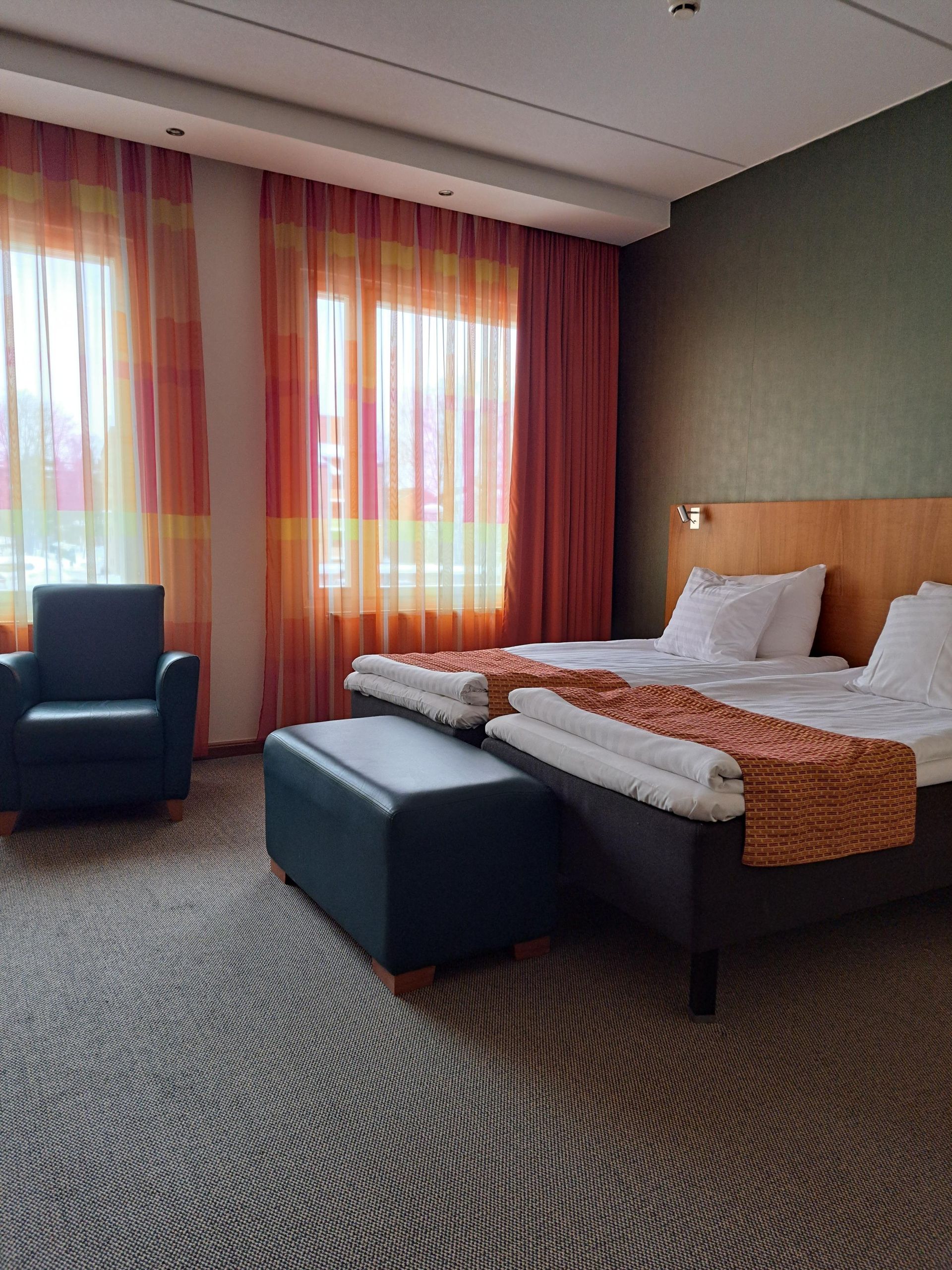 Hotellihuone, jossa on kaksi vierekkäistä erillistä sänkyä, tummansininen nojatuoli, yhteensopiva jalkarahi ja oranssinväriset kuviolliset verhot.