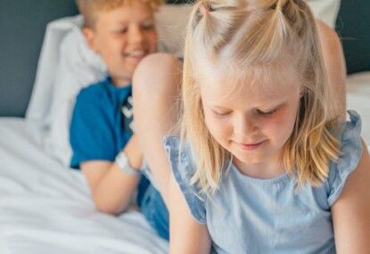 Kaksi lasta istuu sängyllä, toinen etualalla katsoo alas ja toinen taustalla hymyilee.