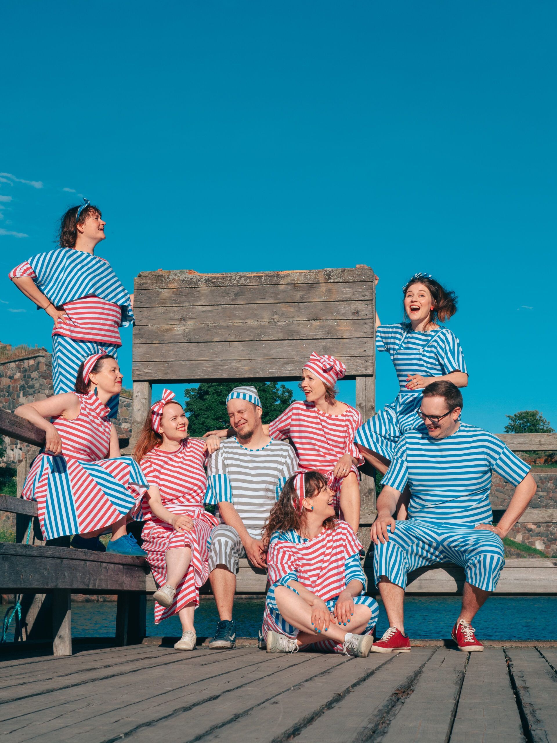 Hattu ja liivi yllään oleva henkilö istuu hymyillen vintage-pinkillä sohvalla sinivihreää, teksturoitua seinää vasten.