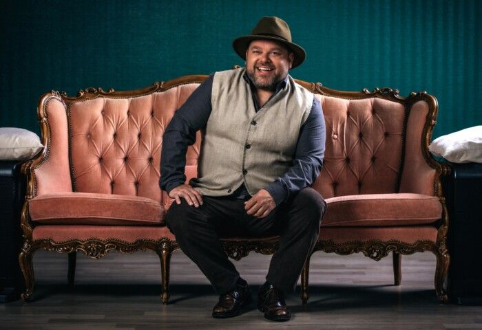 A person wearing a hat and vest smiles while sitting on a pink, tufted velvet couch against a teal-paneled wall.
