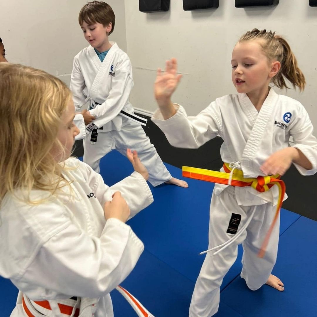 People practicing Brazilian Jiu-Jitsu. One person on the ground, another on top; two others grappling nearby. Black gis, indoor setting.