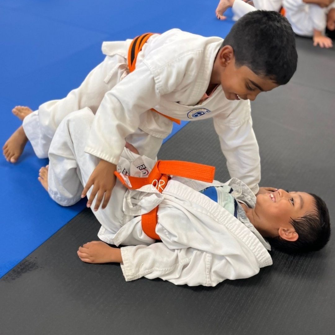 Two young children in blue and black martial arts uniforms grappling on a gym floor, one smiling.