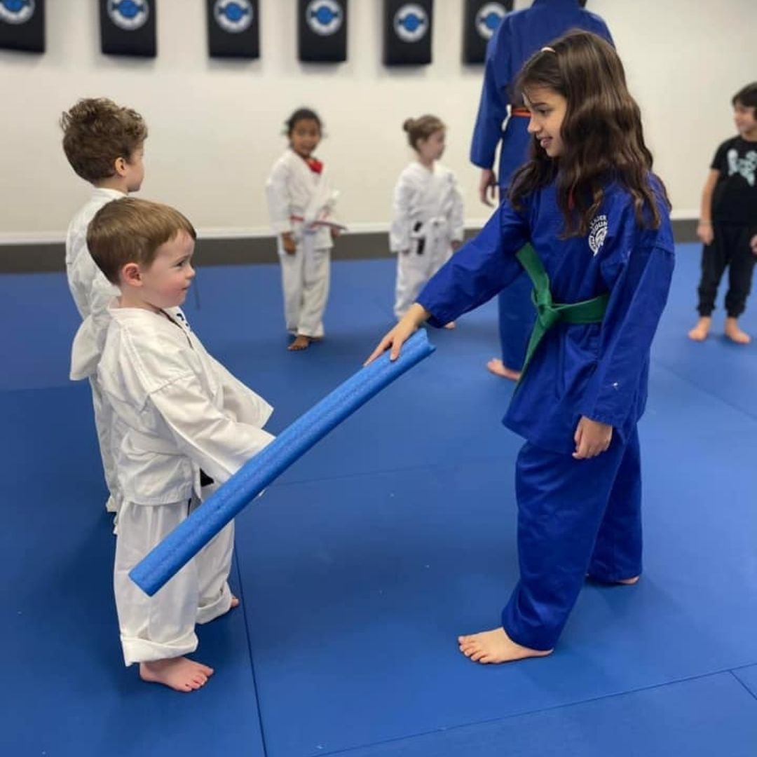 Two children practicing Brazilian Jiu-Jitsu on a mat. One child in white gi has the other in a leg lock. A girl watches.