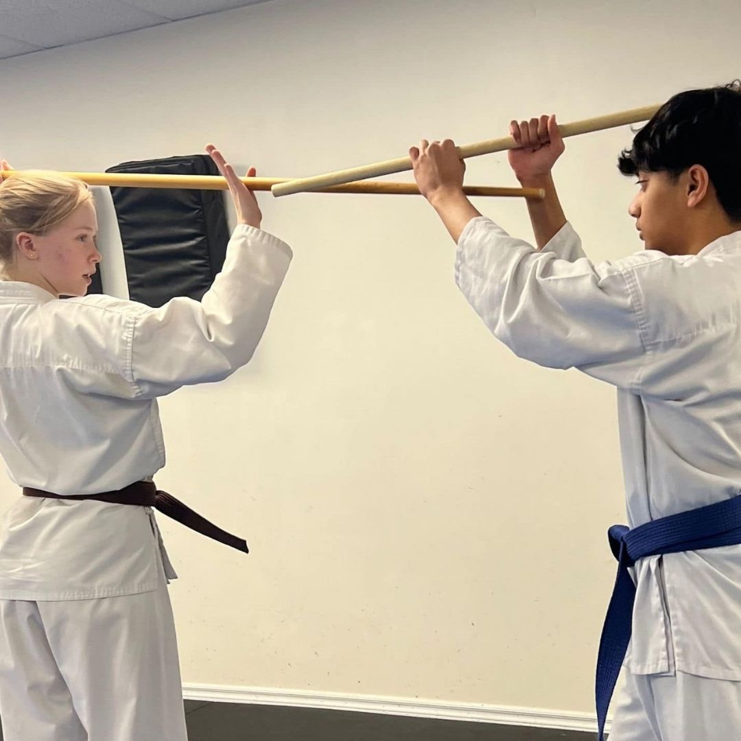 Two men in grappling position on a mat. One embraces the other from behind, both smiling. Gym setting.
