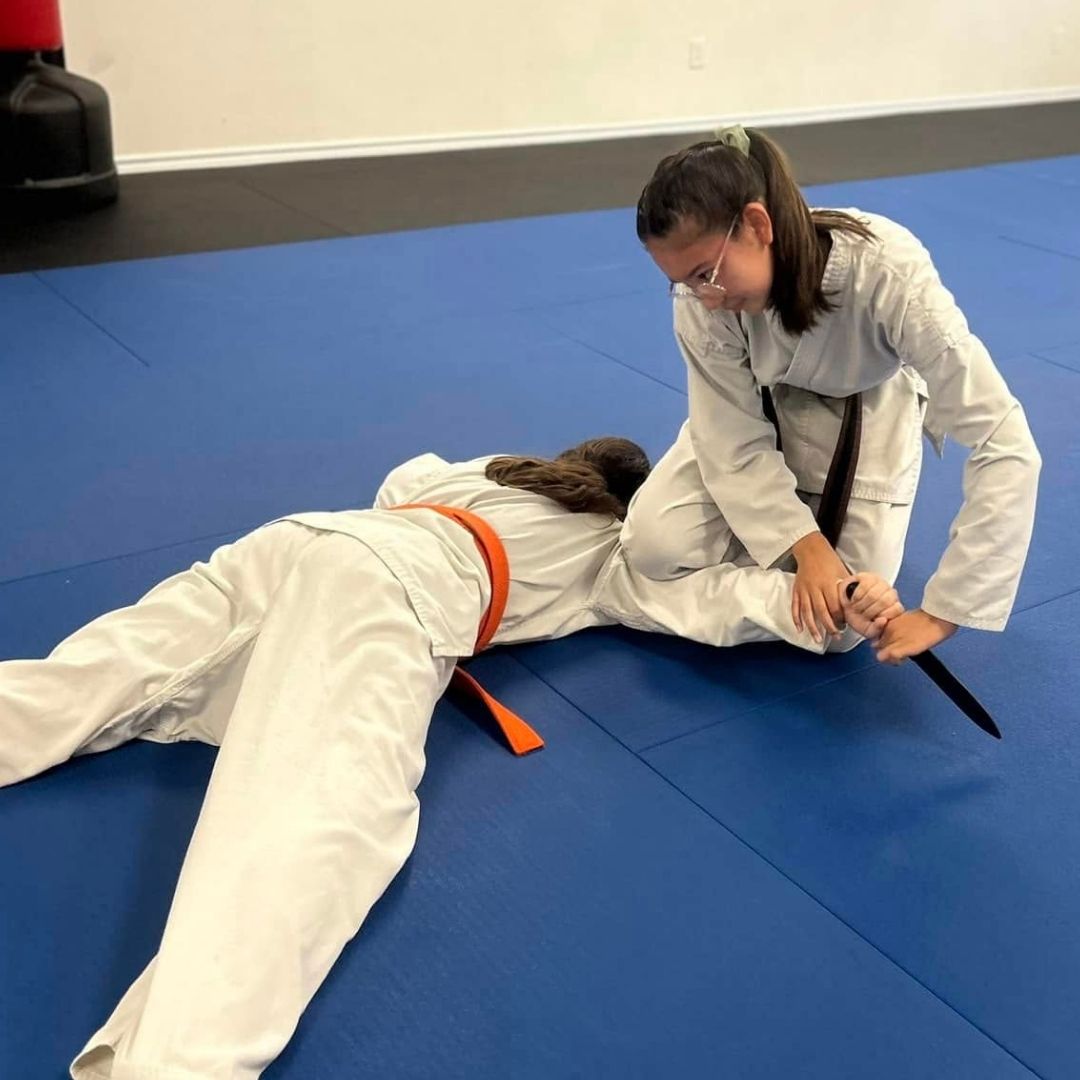 People practicing jiu-jitsu on mats, wearing gis in blue, black, and gray. Active grappling, gym setting.