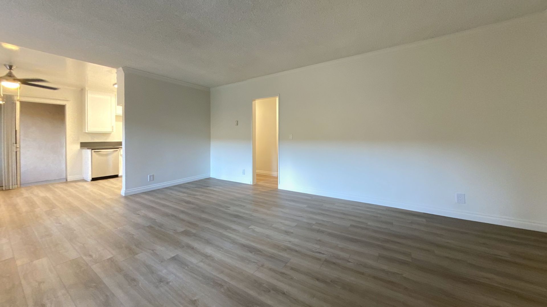 Empty apartment interior with light wood flooring, white walls, and a view into the kitchen and another room.