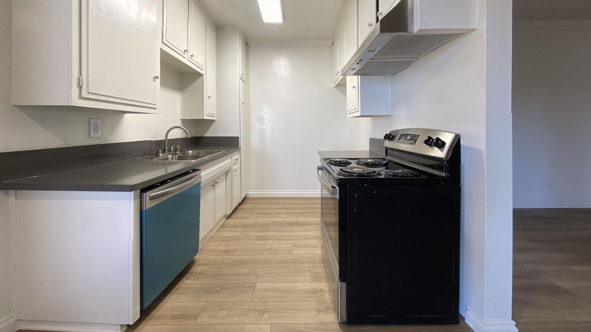 A narrow kitchen with white cabinets, blue dishwasher, black stove, and light wood flooring.