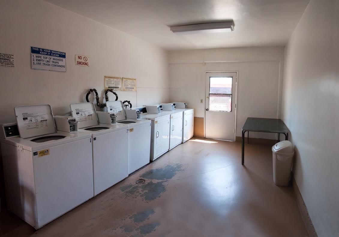 Laundry room with several white washing machines and a door. Pale flooring and walls, and a small table.