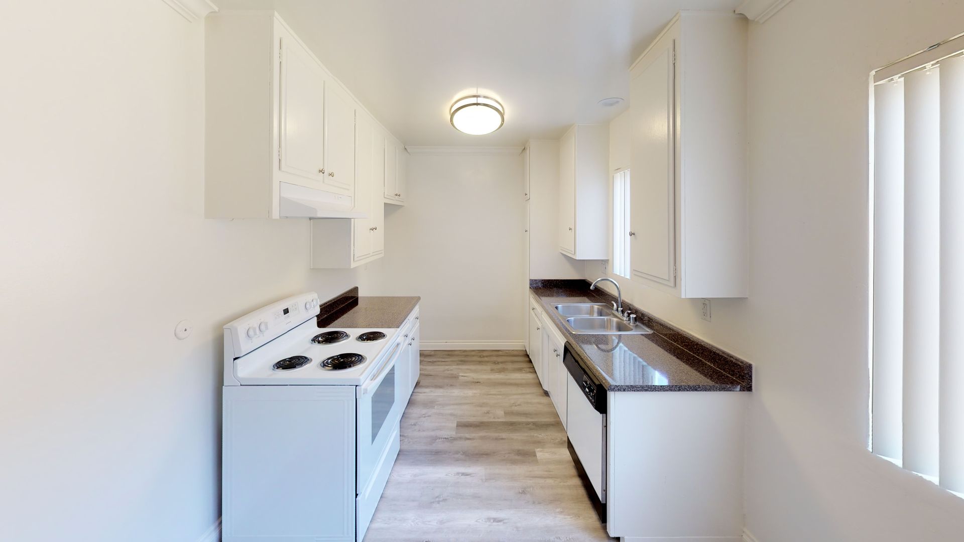 A narrow kitchen with white cabinets, appliances, and a window with blinds.