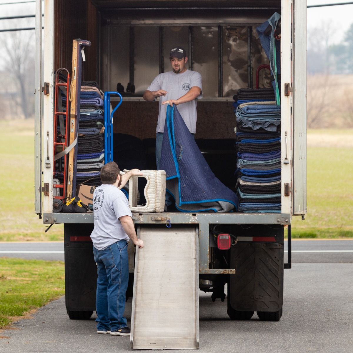 Two movers loading a truck with protective blankets. One stands in the truck, the other on the ramp.