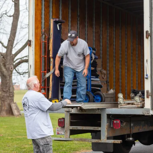 Two movers loading a truck. One stands inside, pushing a dolly; the other outside, assisting on the lift.