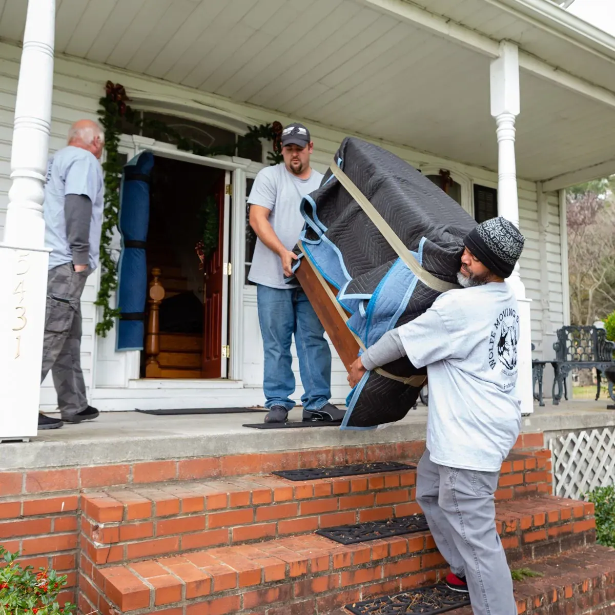 Three people moving a large, wrapped piece of furniture up steps of a house.