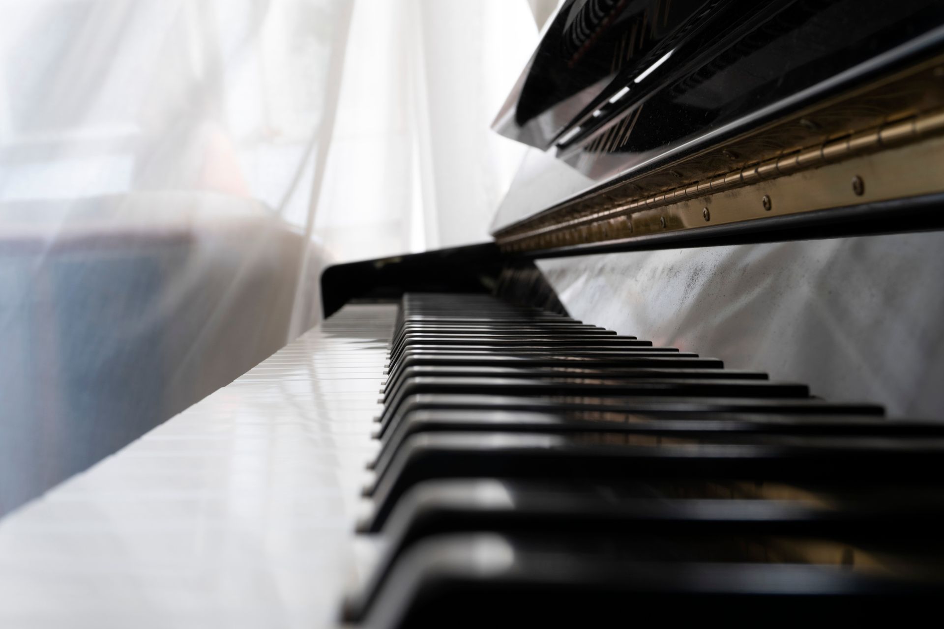 Piano keyboard, close-up. Black and white keys, open lid, gold trim, against white sheer curtain.
