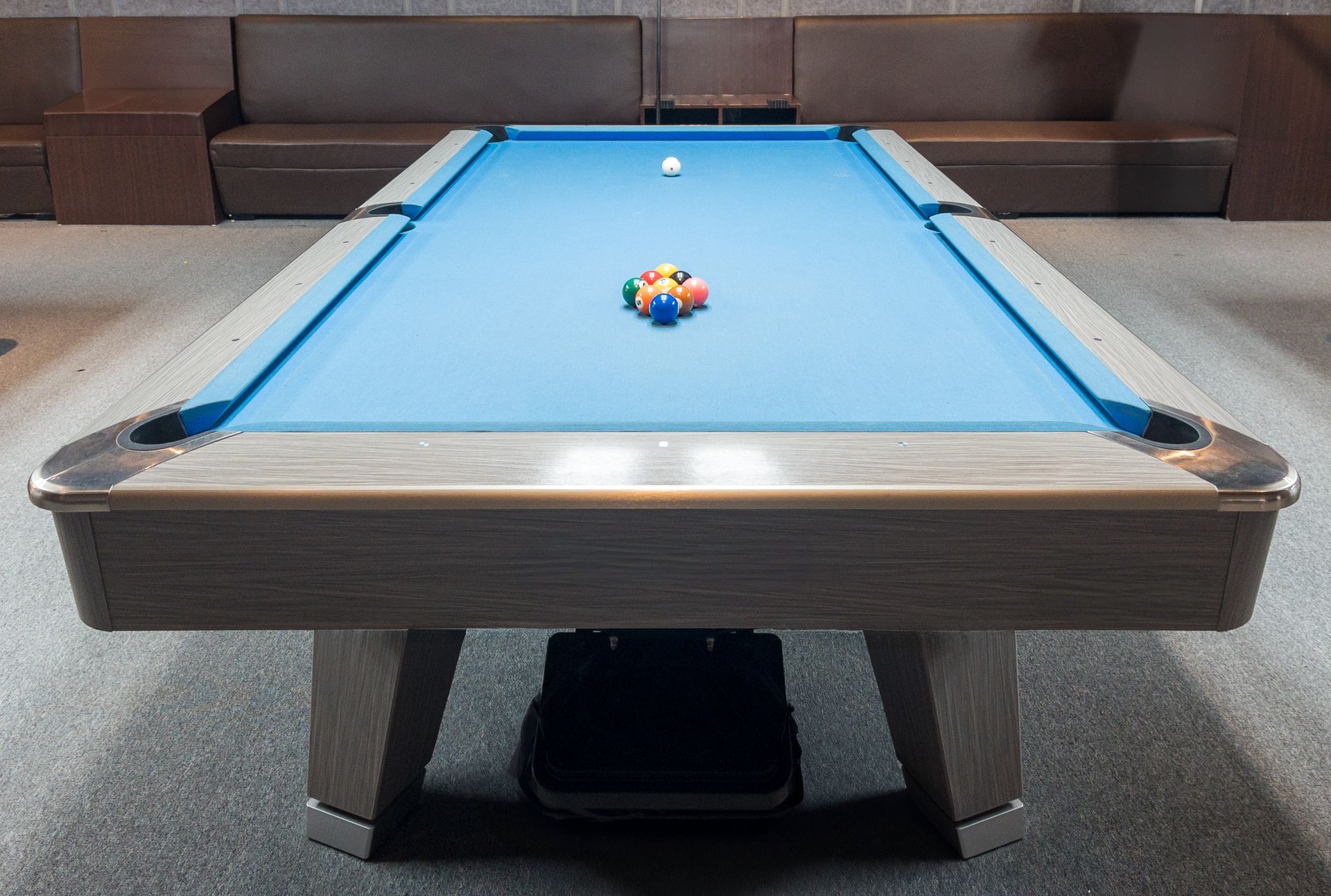 Pool table with balls racked, cue ball at the ready. Blue felt, light wood frame, brown seating in background.
