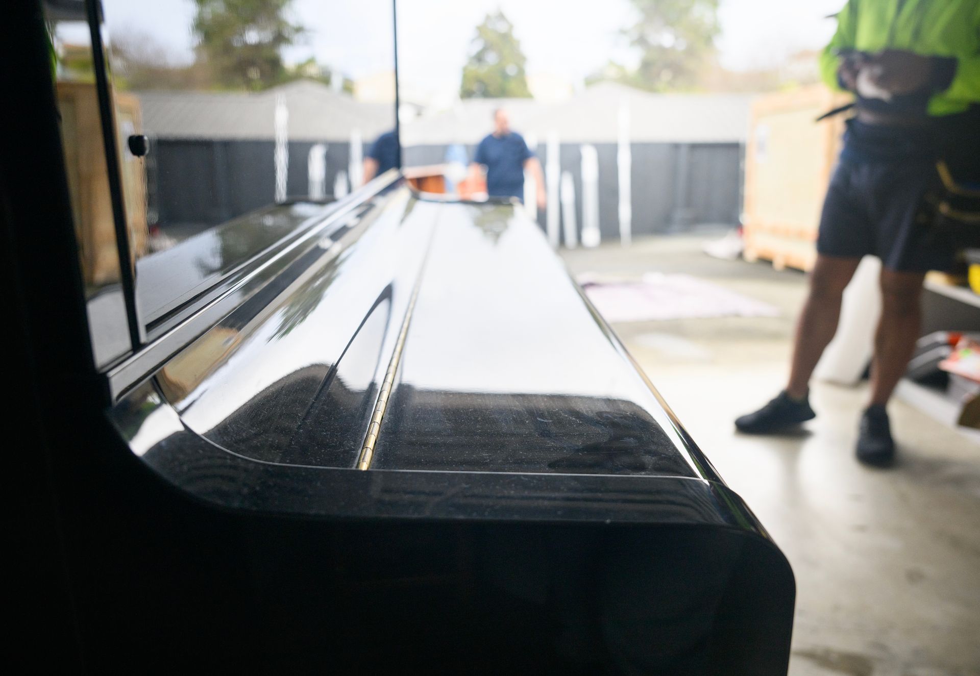 Close-up of a shiny black piano with two people blurred in the background, likely movers in a garage.
