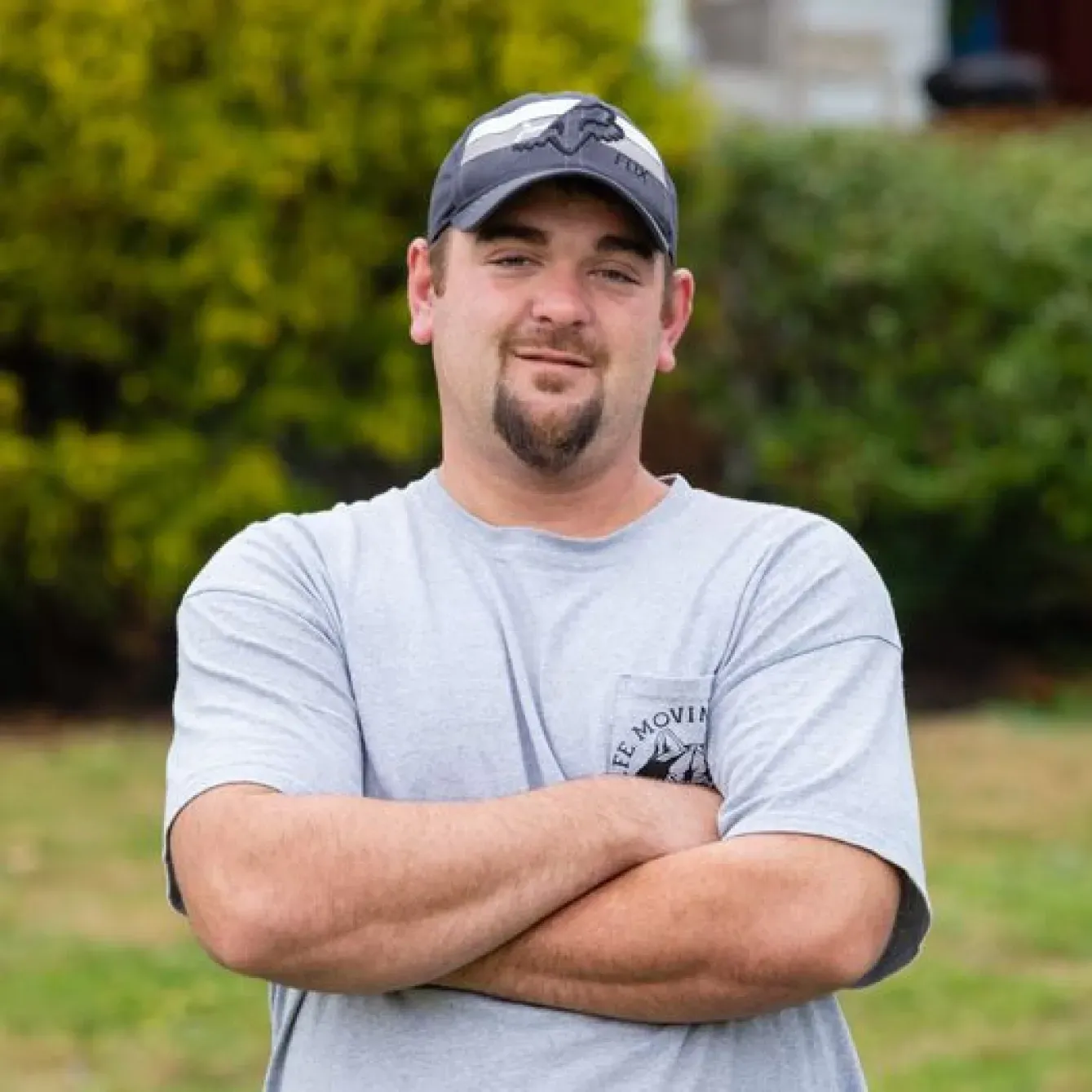 Man with arms crossed, wearing a cap and gray t-shirt, standing outdoors in front of green foliage.