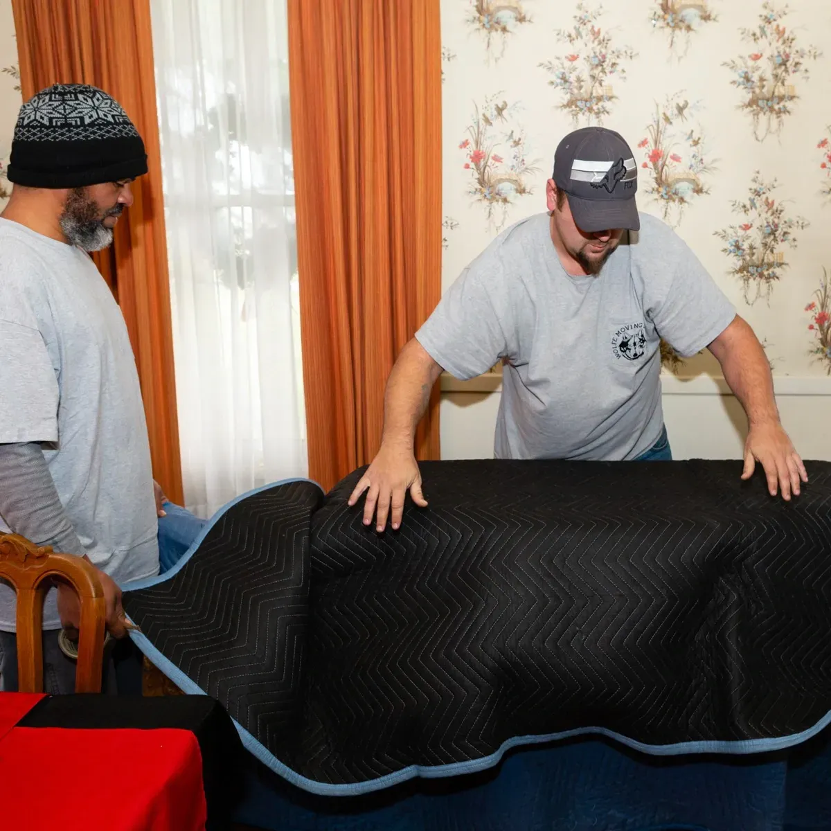 Two movers wrapping furniture in a room with floral wallpaper and orange curtains.