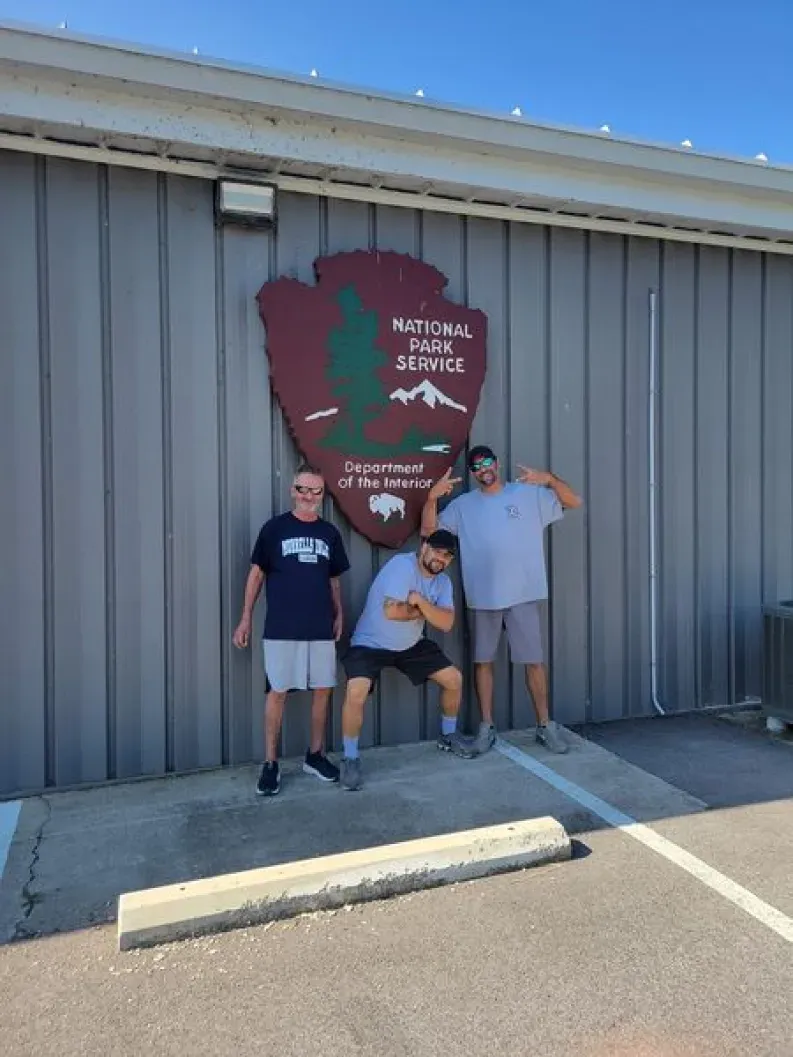 Three people posing in front of a National Park Service sign on a gray building, sunny day.