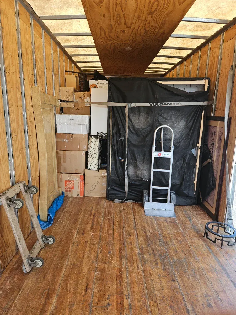 Interior of a moving truck filled with boxes, furniture, and a hand truck. Wooden floor and walls.