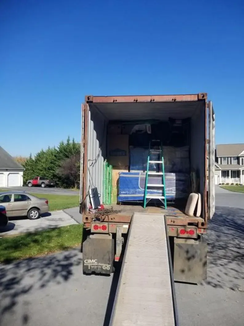 A truck's cargo area filled with various items; a ramp extends to the ground under a blue sky.