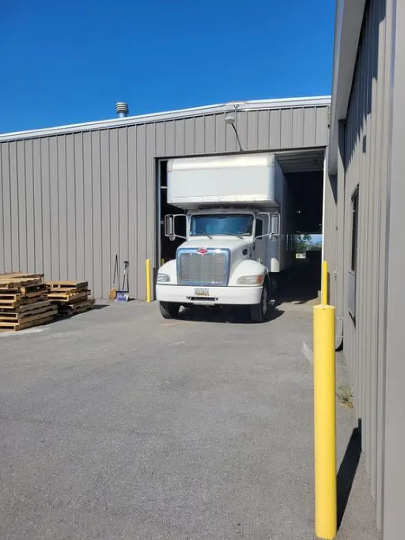 A white box truck loading at a warehouse with open bay doors under a bright blue sky.