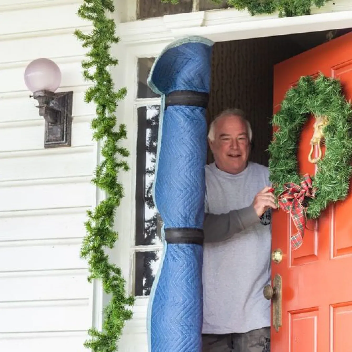 Man holds rolled-up blue moving blanket in a doorway.  A Christmas wreath adorns the orange door. Green garland hangs.