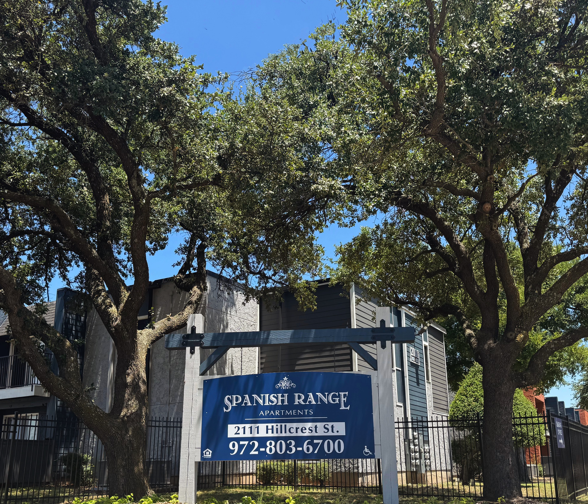 Sign for Spanish Range Apartments: 2111 Miller St. 972-803-6700. Two trees frame the sign and building.
