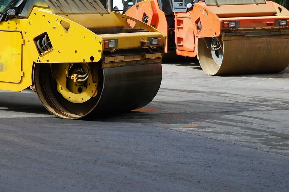 Yellow road roller compacting fresh asphalt, orange roller in background.