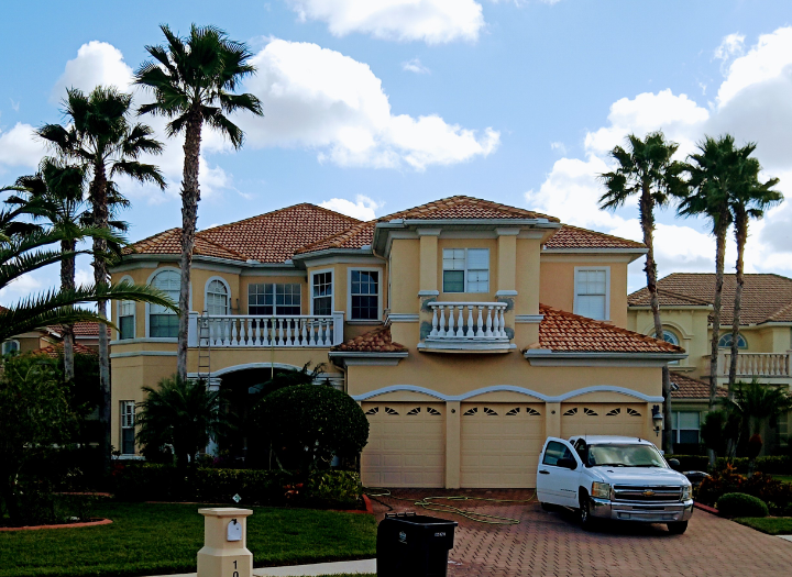 Two-story beige house with red-tile roof, three-car garage, and a white truck in the driveway; palm trees and blue sky.
