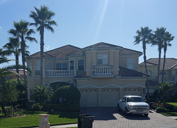 Two-story beige house with red-tiled roof and three-car garage; palm trees; a silver pickup truck parked in front.