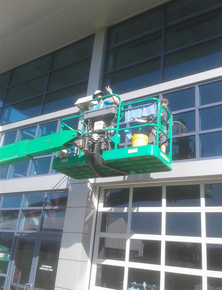 A person in a lift cleaning the exterior windows of a building. Green lift arm and bucket, shiny windows.