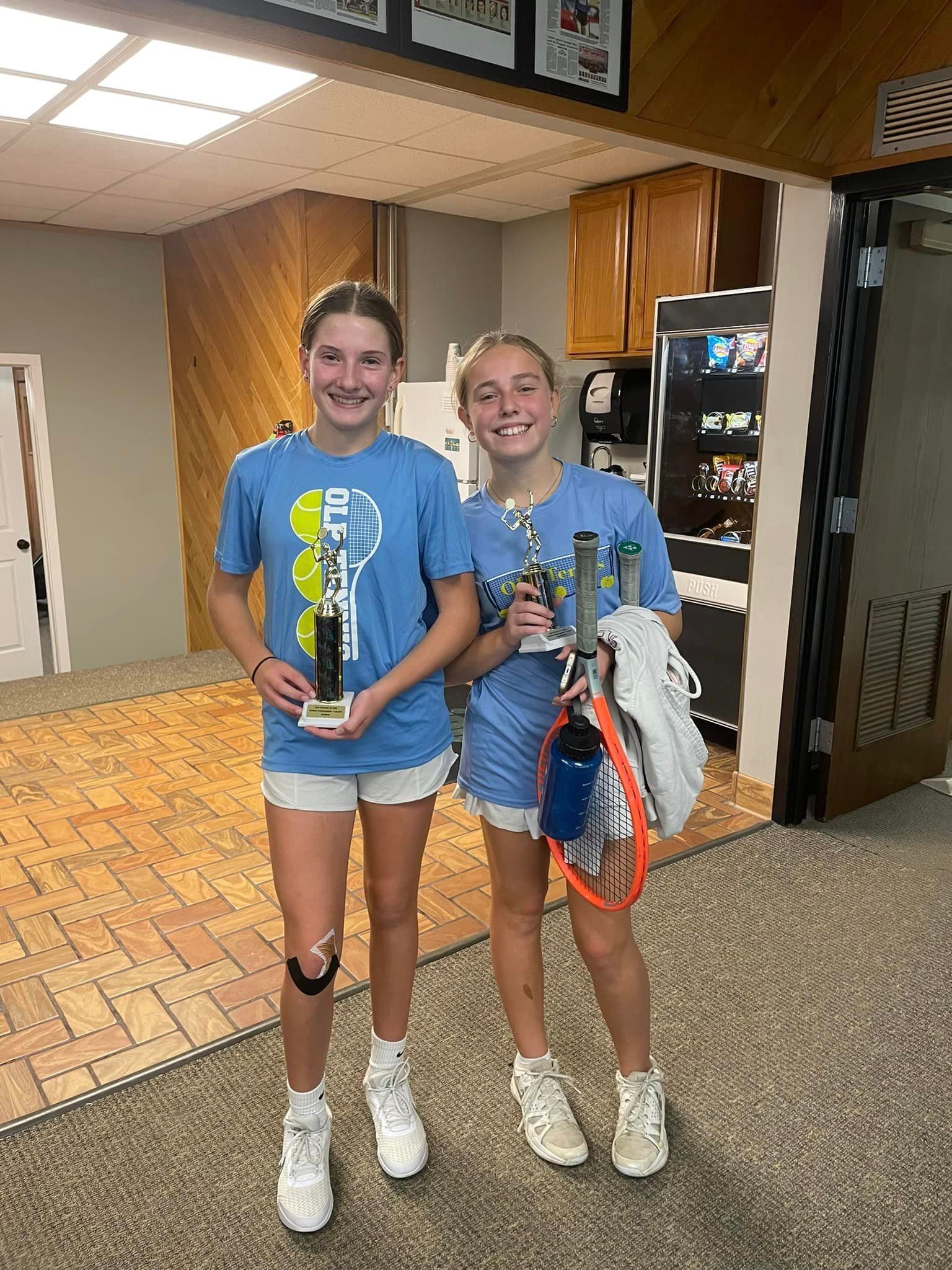 Two girls holding tennis rackets and trophies in a room