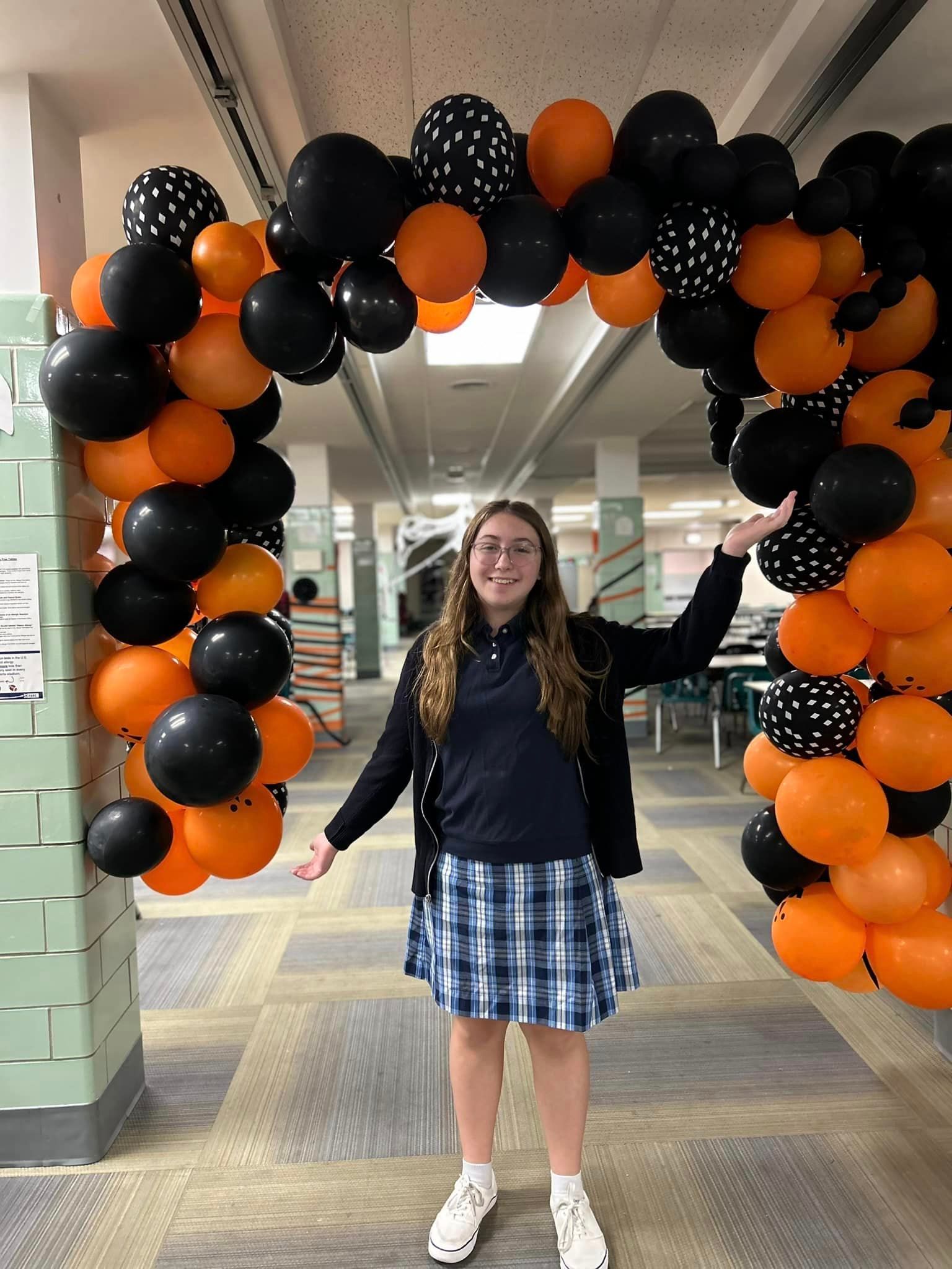 A girl in a plaid skirt is standing in a hallway decorated with black and orange balloons
