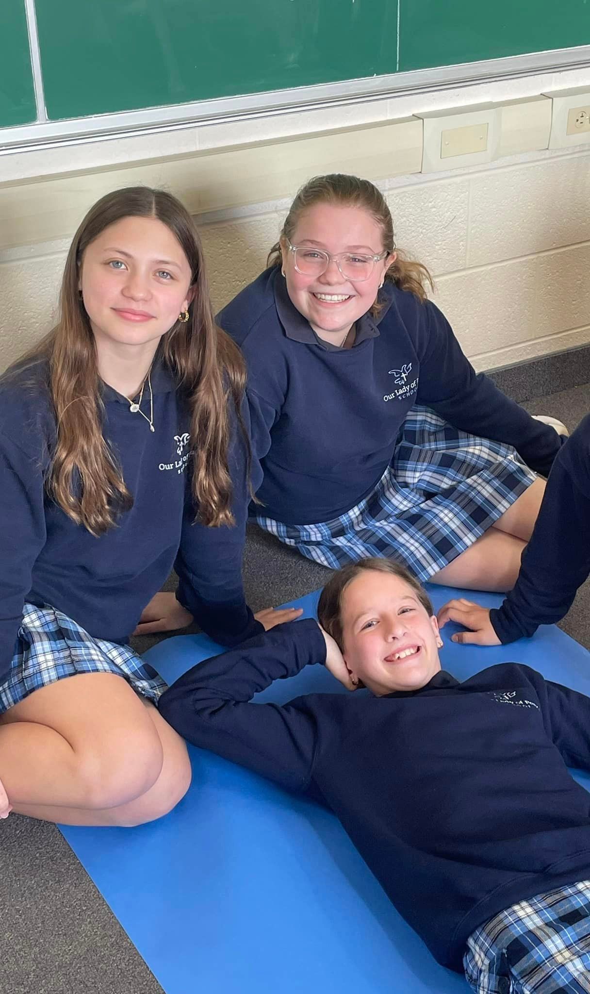 Three girls in school uniforms are sitting on a yoga mat