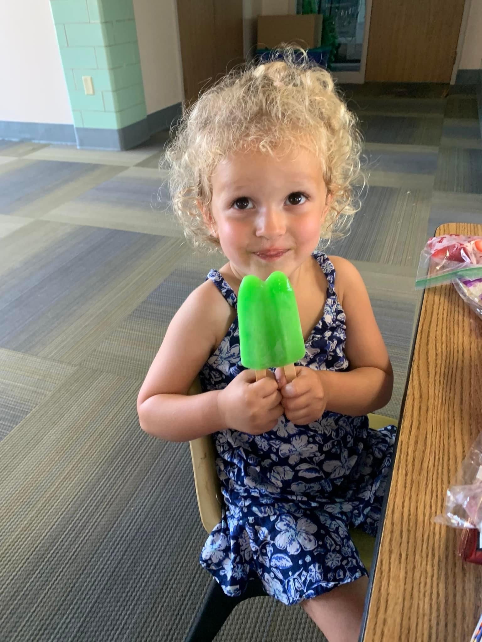 A little girl is sitting at a table holding a green popsicle