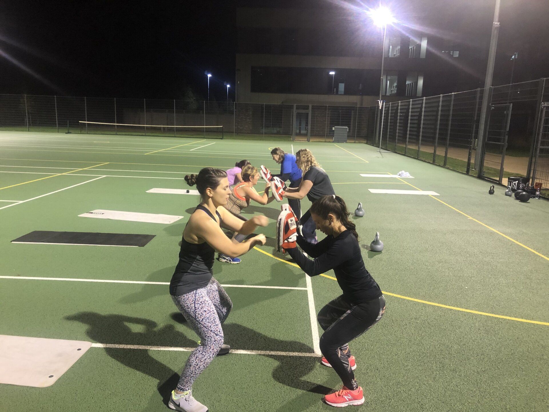 A group of women are doing squats on a tennis court at night.
