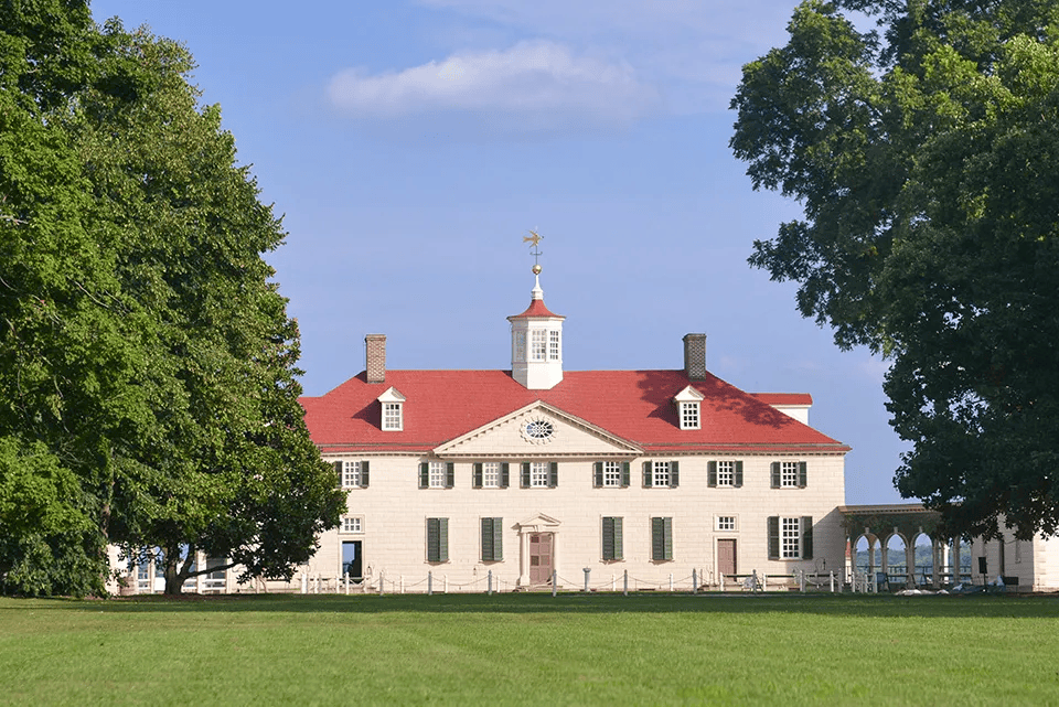 Mount Vernon estate with red roof, white facade, and cupola against a blue sky, framed by trees.