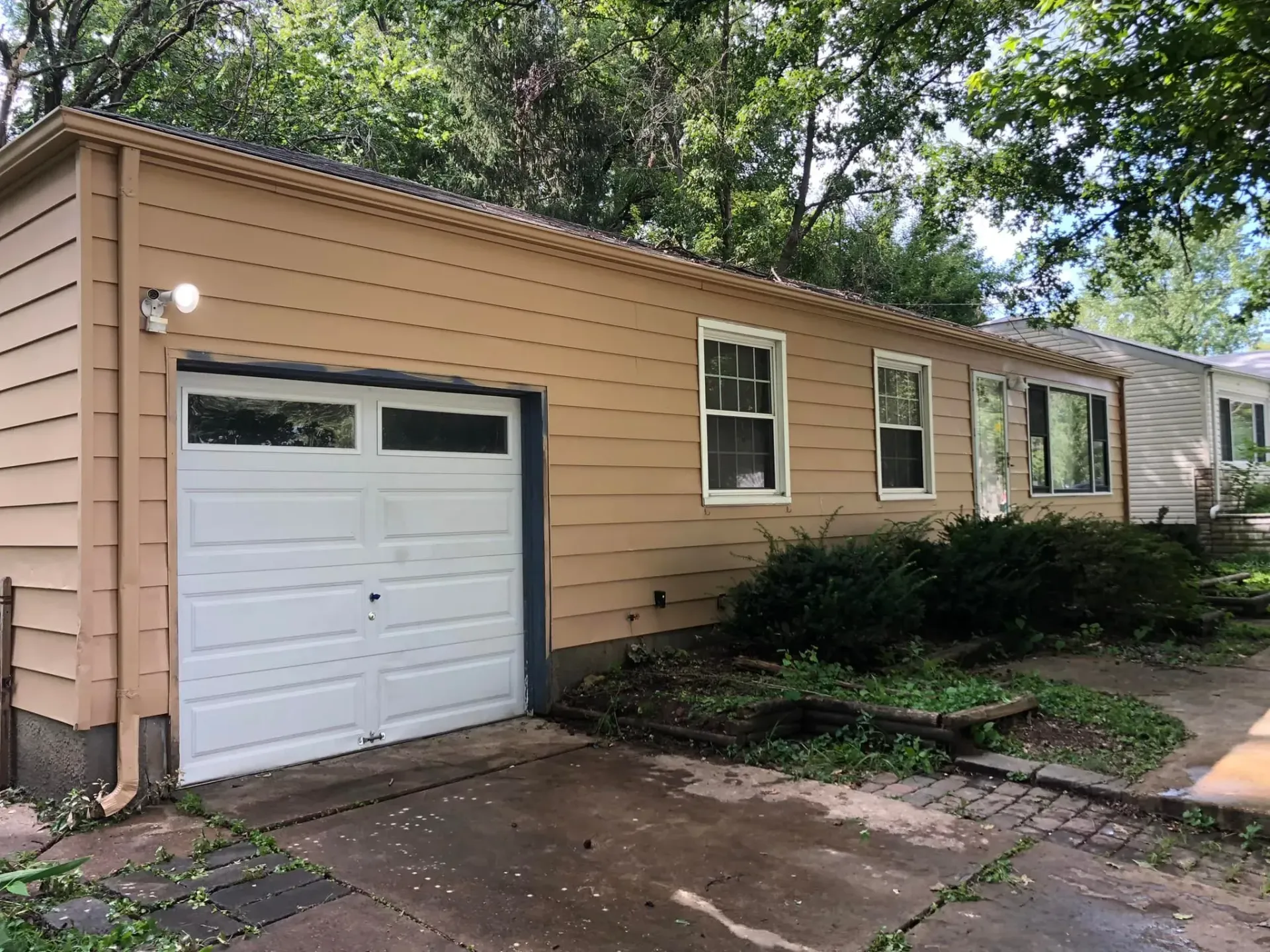 Beige house with a white garage door, windows, and overgrown bushes.