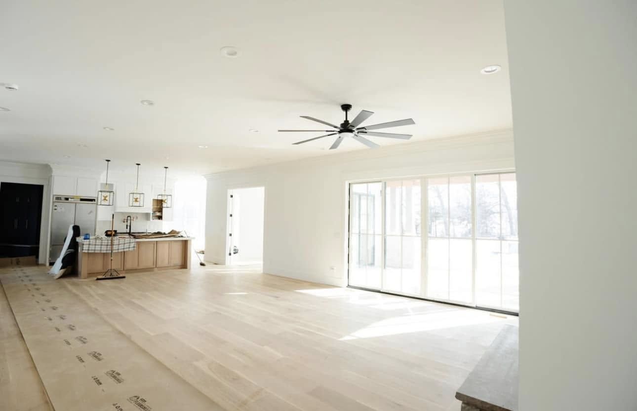 Bright, empty living space with light wood floors, white walls, and a large ceiling fan.