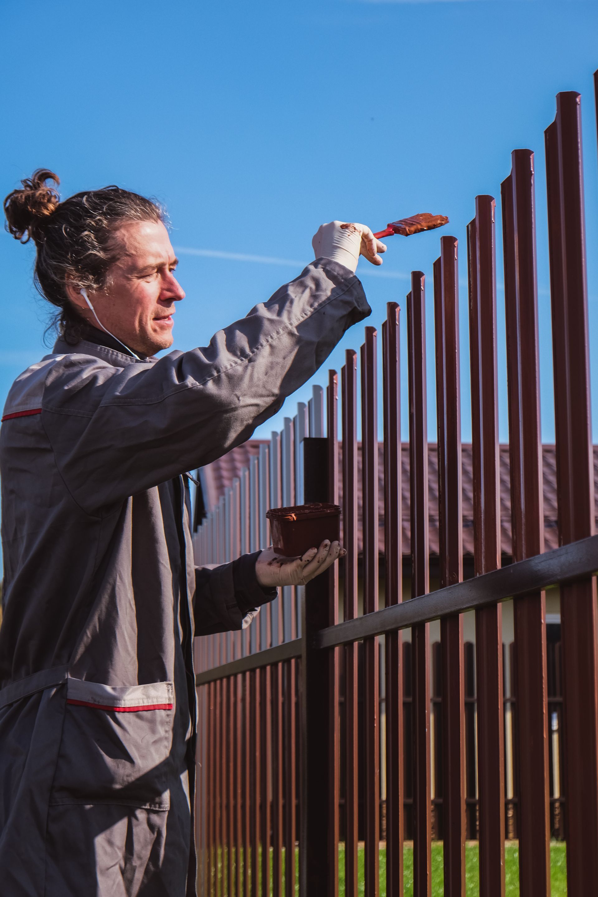 Person paints a brown metal fence outdoors on a sunny day, holding a paintbrush and paint. Person paints a brown metal fence outdoors on a sunny day, holding a paintbrush and paint.