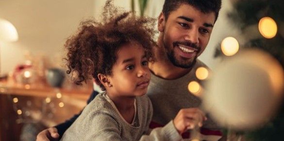 A father and child decorate a Christmas tree together. The father smiles, and the child looks intently.