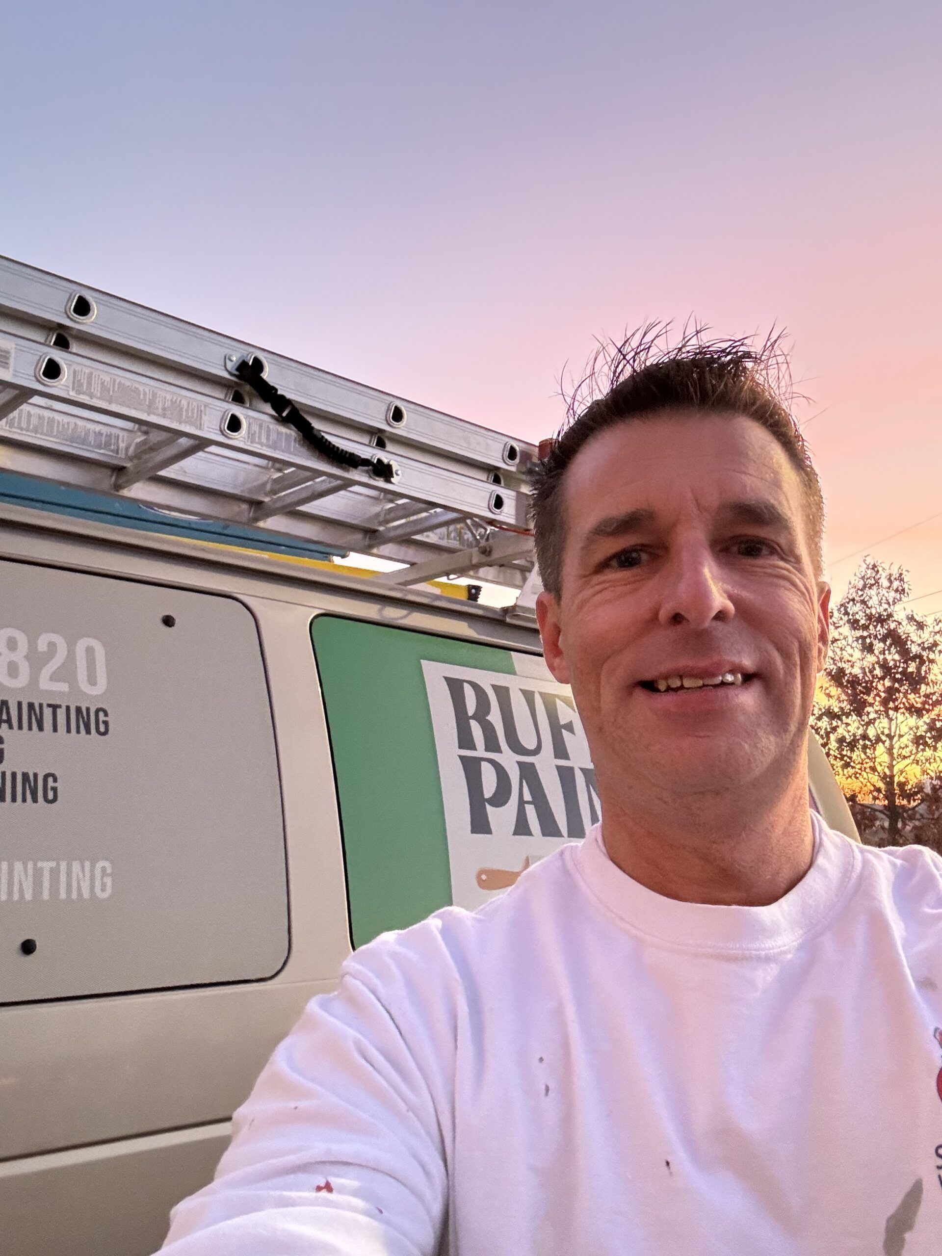 Man in white shirt smiling next to a van with a ladder. Sunset in the background. Van says