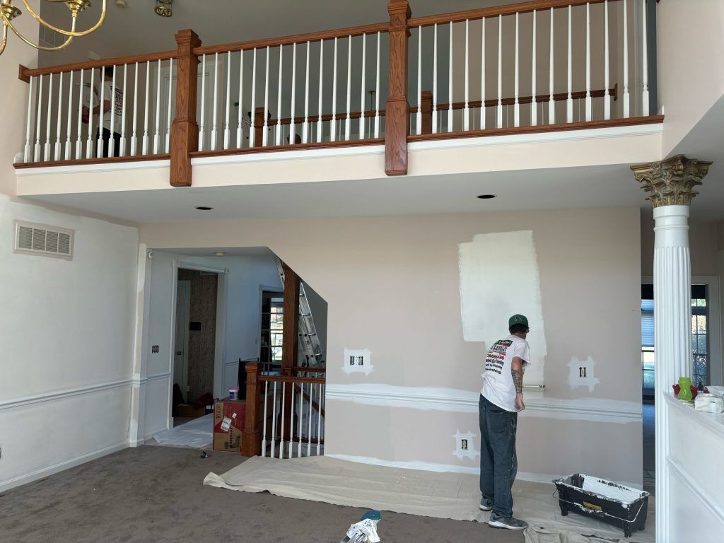 Person painting a wall inside a house. Beige walls, brown trim, carpeted floor, and a balcony overhead.