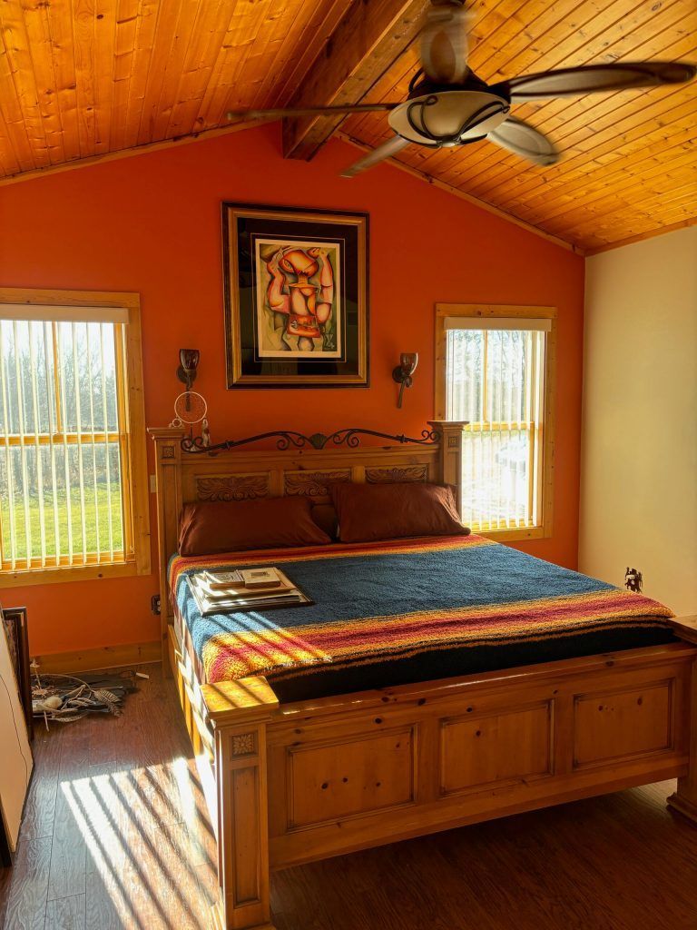 Bedroom with orange walls and wooden ceiling, bed, windows, and artwork.